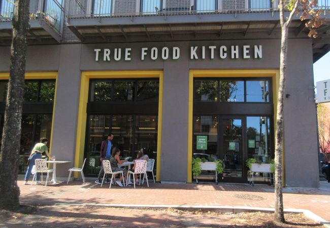 Entrance of True Food Kitchen with outdoor seating, featuring a group of people dining and trees lining the sidewalk.