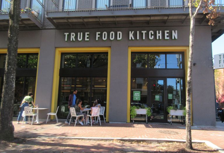 Entrance of True Food Kitchen with outdoor seating, featuring a group of people dining and trees lining the sidewalk.