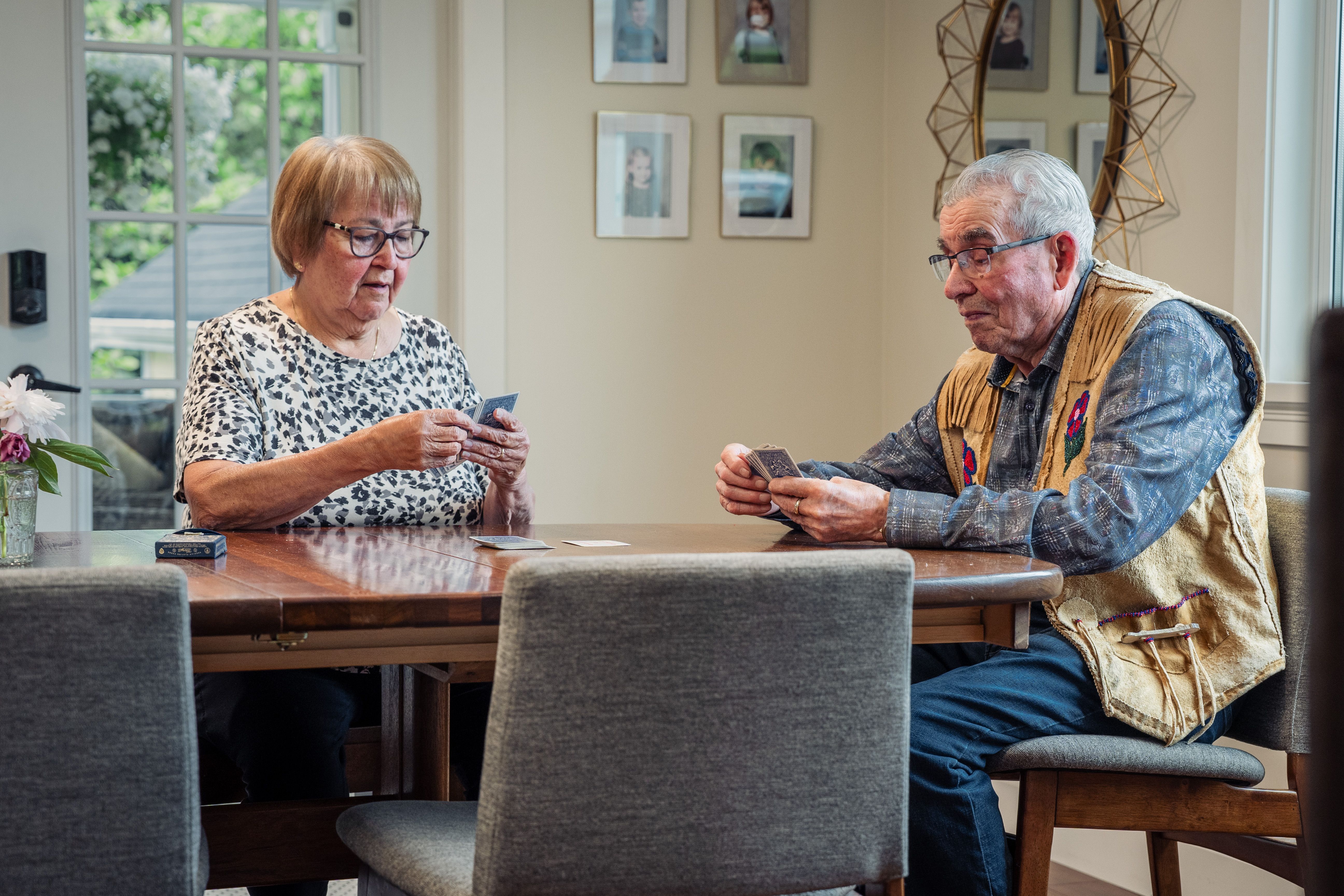 Two elderly people sitting at a wooden table playing cards in a room with framed photos on the wall.