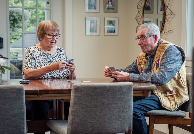 Two elderly people sitting at a wooden table playing cards in a room with framed photos on the wall.