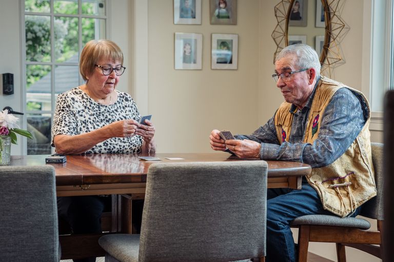 Two elderly people sitting at a wooden table playing cards in a room with framed photos on the wall.