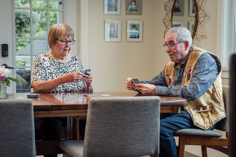 Two elderly people sitting at a wooden table playing cards in a room with framed photos on the wall.