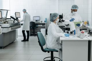 Scientists in protective gear work in a lab; one uses a microscope while another takes notes.