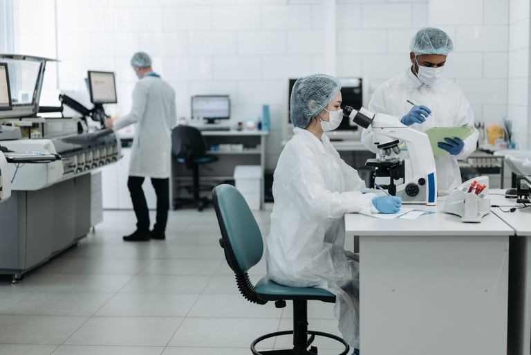 Scientists in protective gear work in a lab; one uses a microscope while another takes notes.