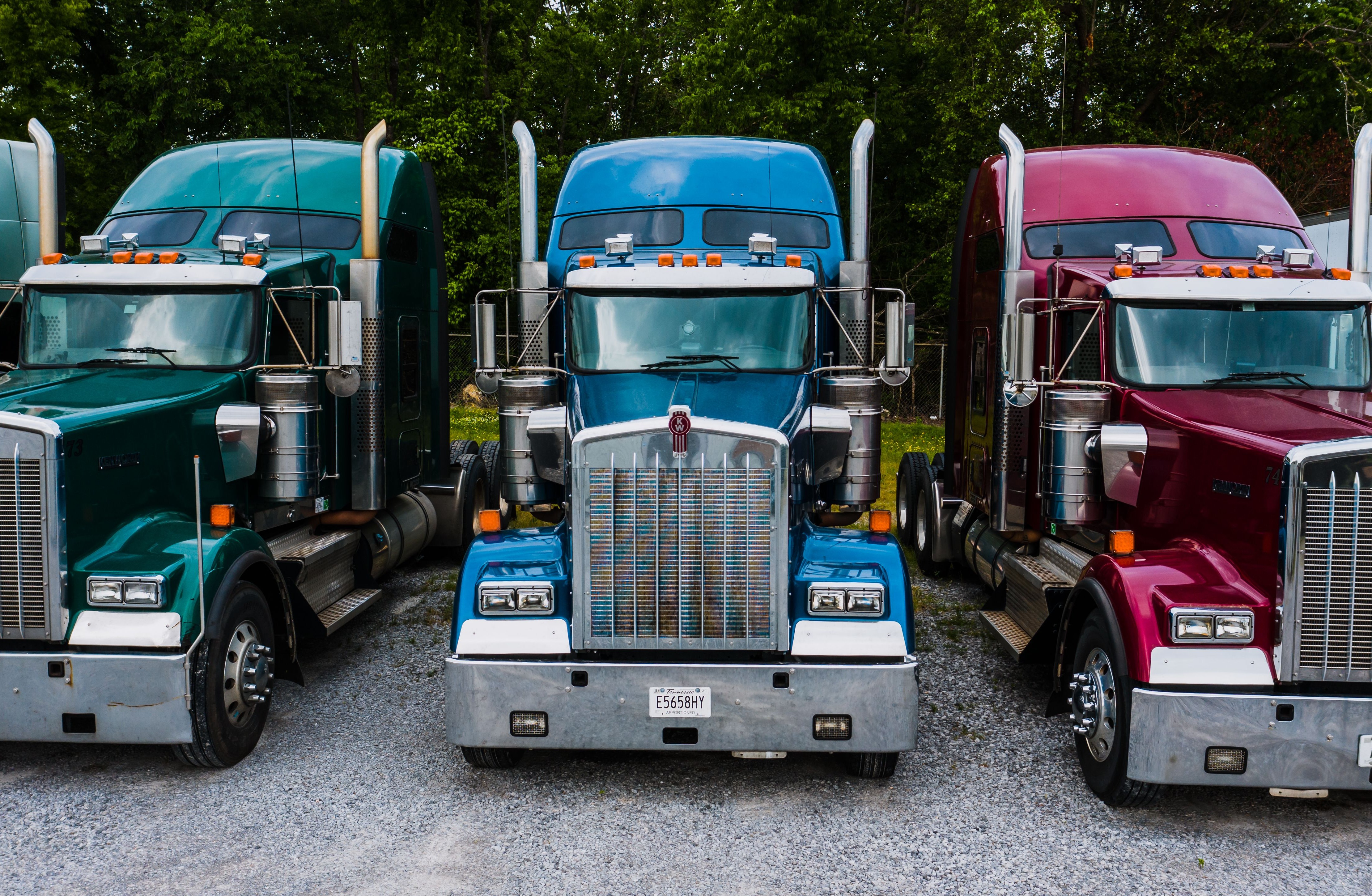 Three colorful semi-trucks, green, blue, and red, parked side by side on a gravel lot with trees in the background.