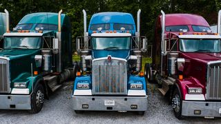 Three colorful semi-trucks, green, blue, and red, parked side by side on a gravel lot with trees in the background.