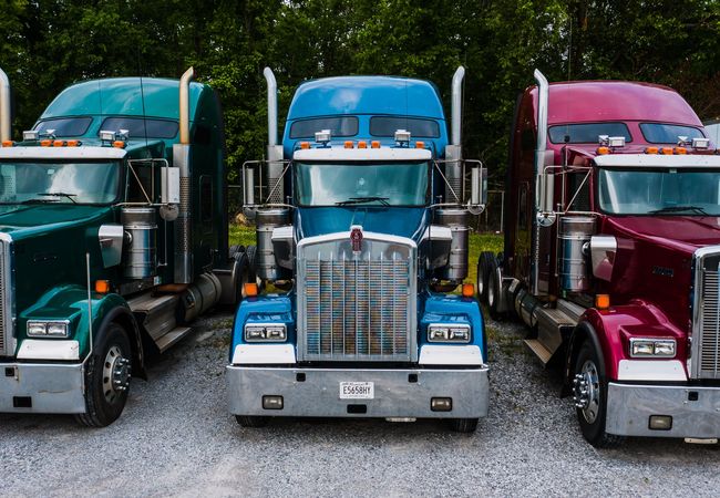 Three colorful semi-trucks, green, blue, and red, parked side by side on a gravel lot with trees in the background.