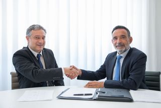 Two men in suits shaking hands over a table with documents and a pen, smiling, in a well-lit office setting.