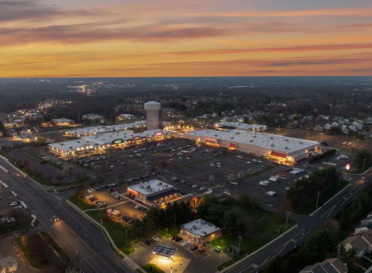 Aerial view of a shopping center at dusk, with illuminated parking lots, surrounding roads, and suburban housing in the background under a colorful sky.