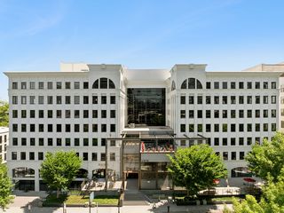 A large, symmetrical white office building with numerous windows, featuring a central glass entrance and rooftop seating, surrounded by trees.
