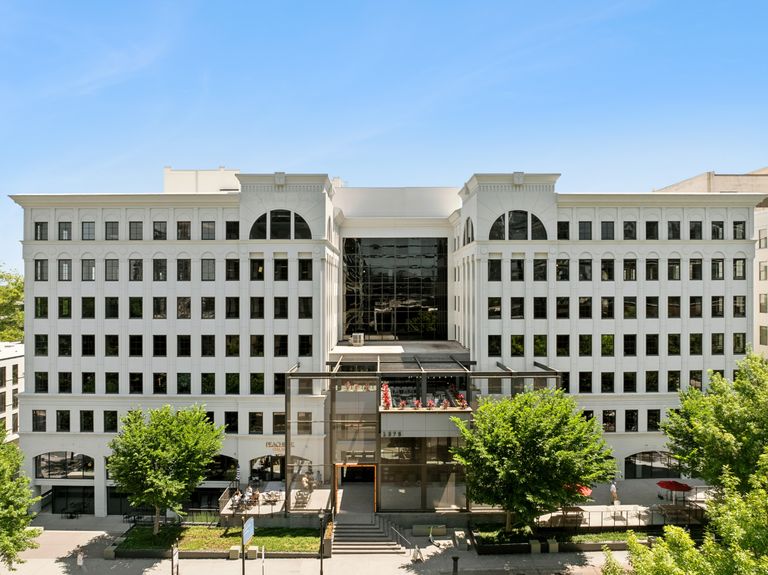 A large, symmetrical white office building with numerous windows, featuring a central glass entrance and rooftop seating, surrounded by trees.