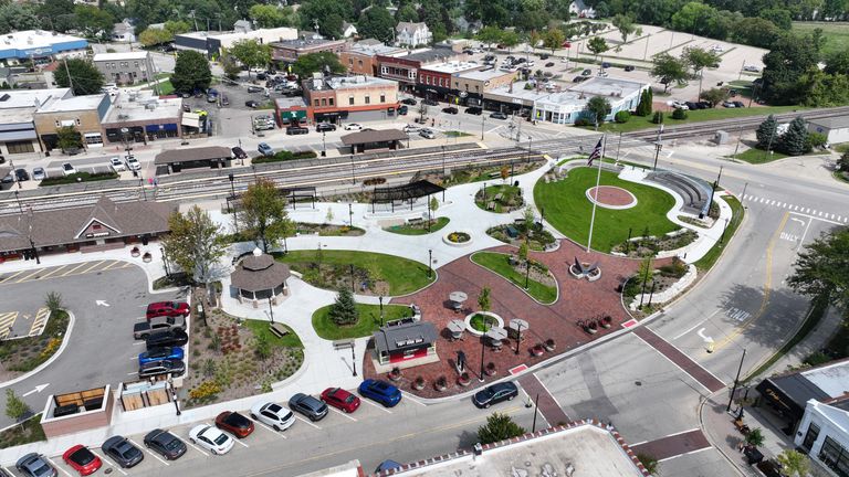 Aerial view of a small urban park with pathways, a gazebo, grassy areas, benches, a circular stage, and adjacent parking and railway tracks.
