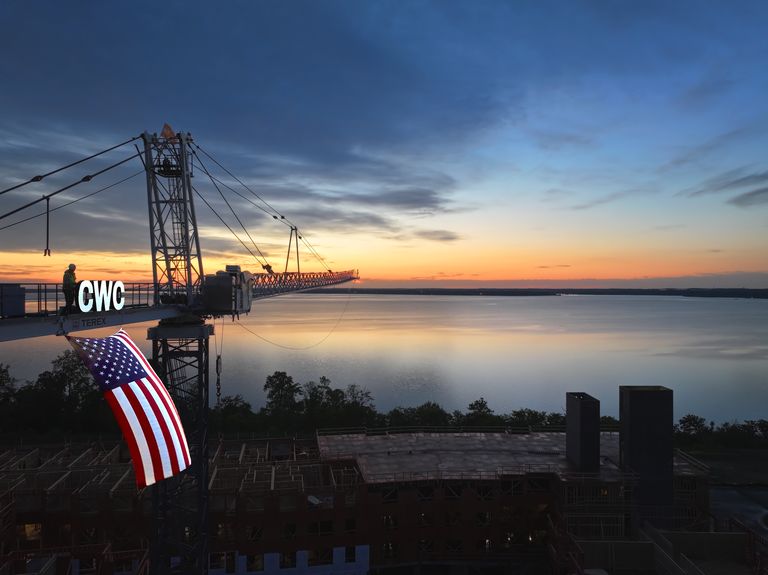 A construction crane silhouetted against a sunrise over a calm body of water, with an American flag hanging from it.