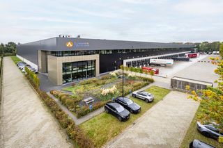 Modern warehouse building with a logo, surrounded by parked cars and greenery, with loading docks and trucks in the background.