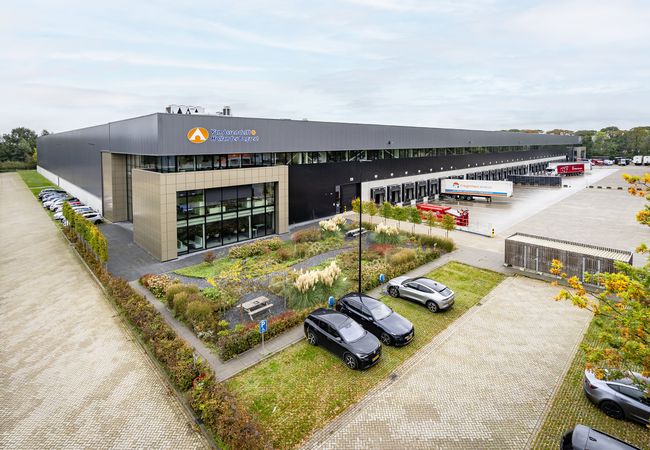 Modern warehouse building with a logo, surrounded by parked cars and greenery, with loading docks and trucks in the background.