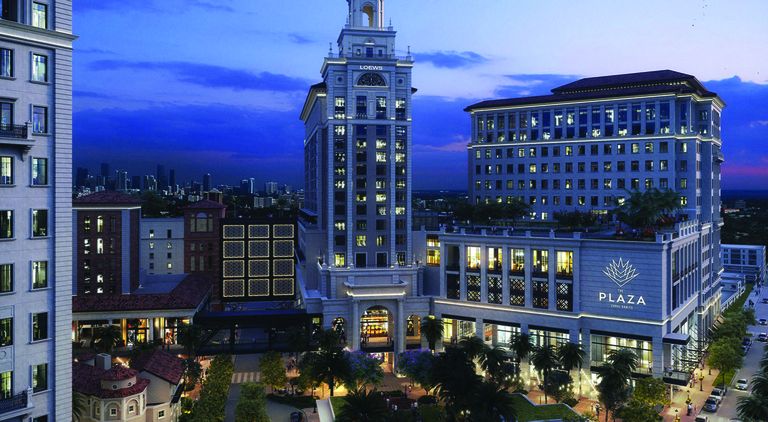 Illuminated plaza building and tower at dusk, surrounded by trees and nearby cityscape under a deep blue sky.