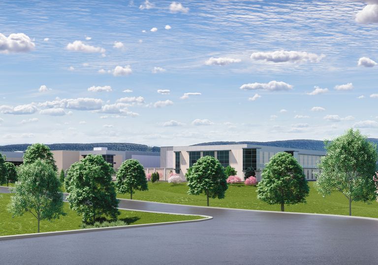 Wide view of a modern building complex surrounded by grass, trees, and a clear blue sky with scattered clouds and distant hills.