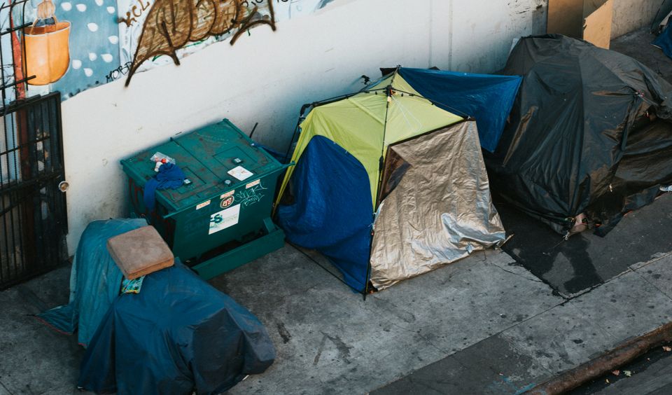 Tents and a dumpster line a city sidewalk beneath a mural on a wall, depicting various scenes and graffiti.