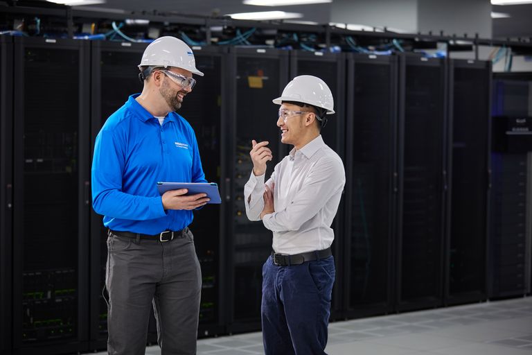 Two men wearing hard hats and safety glasses chat in a server room. One holds a tablet, both smiling, with racks of servers behind them.