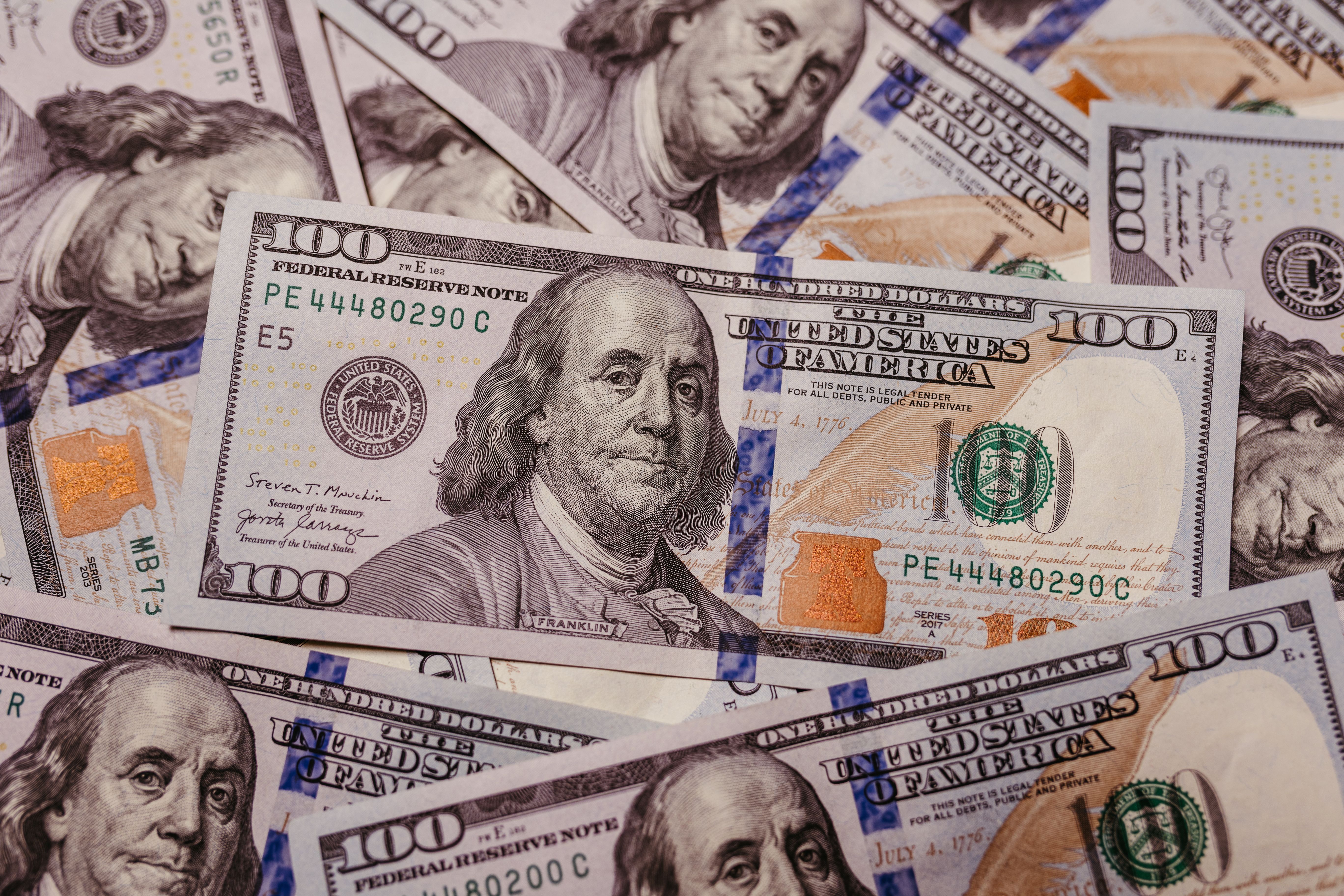 A close-up of multiple U.S. one hundred dollar bills featuring Benjamin Franklin, with visible security features and serial numbers.