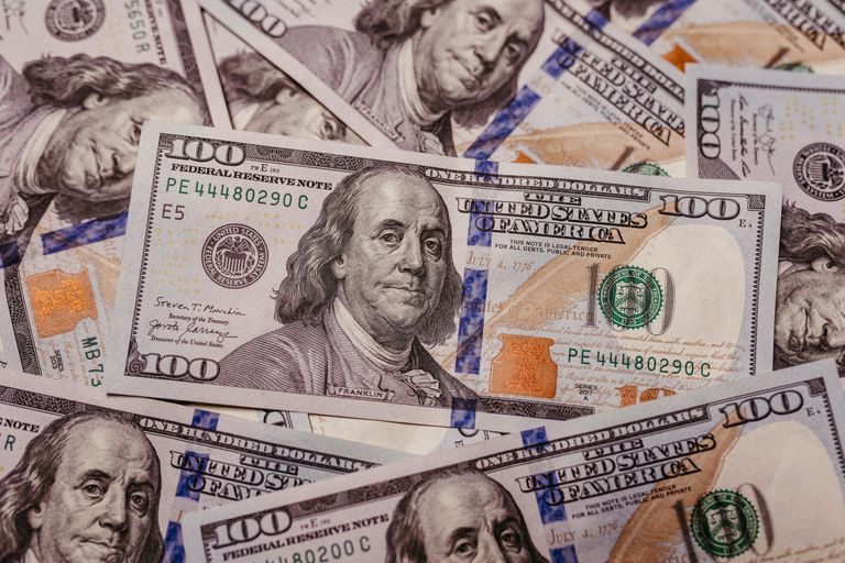 A close-up of multiple U.S. one hundred dollar bills featuring Benjamin Franklin, with visible security features and serial numbers.