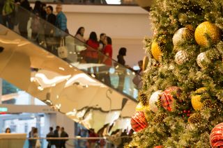 A decorated Christmas tree with large ornaments stands beside an escalator filled with people in a busy shopping mall.