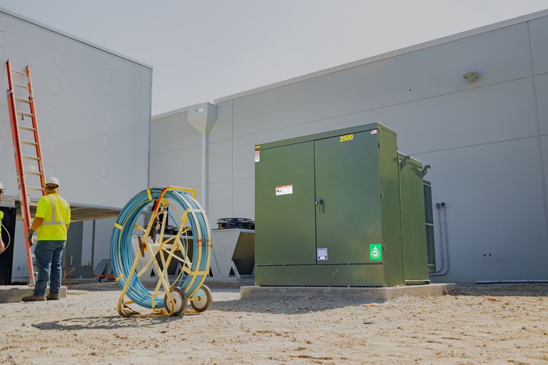 A construction worker stands beside a large green electric transformer and a roll of blue cable outside a gray industrial building.