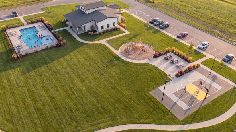 Aerial view of a community park with a swimming pool, playground, basketball court, and picnic area surrounded by green lawns and parking spaces.