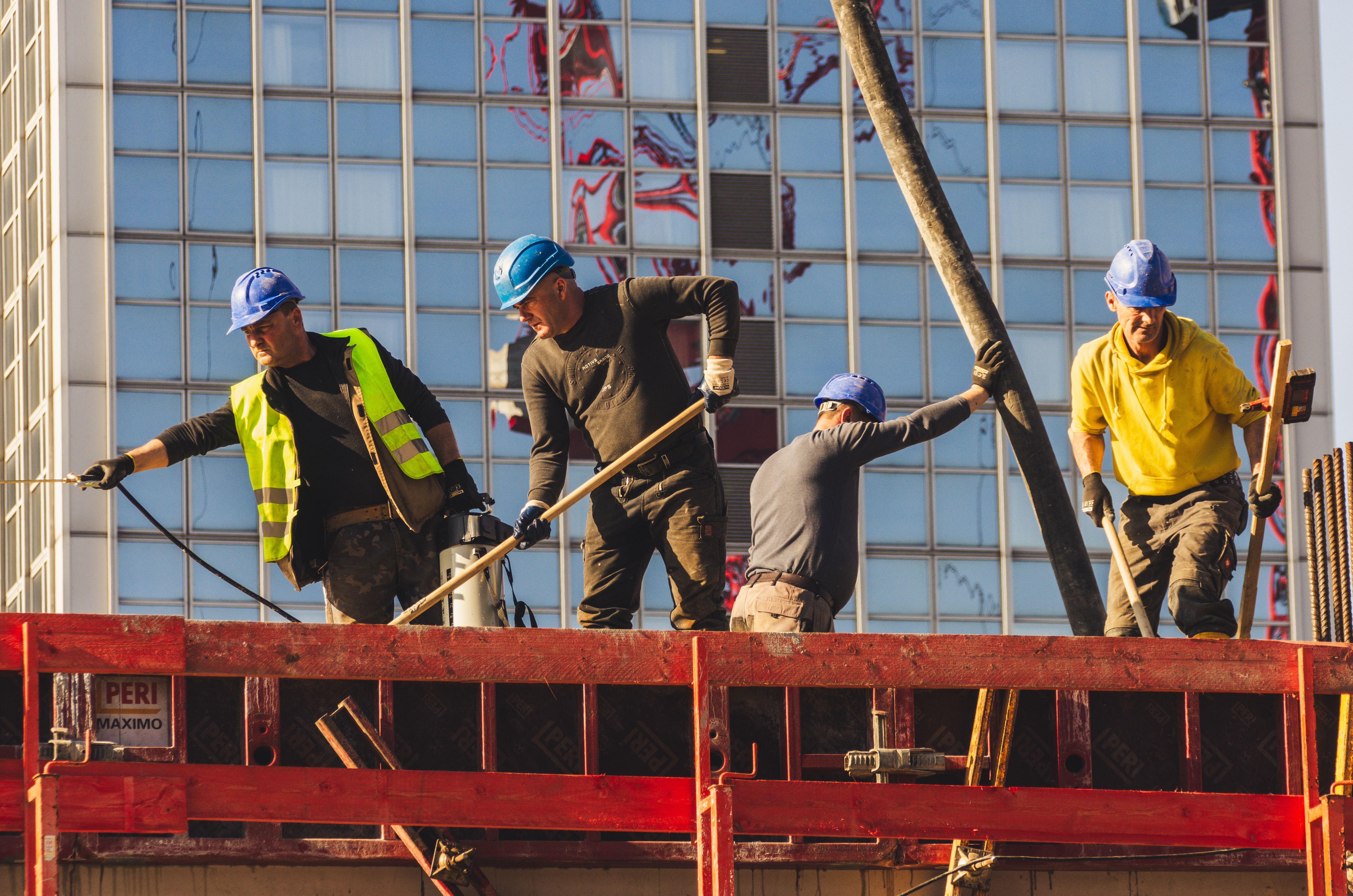 Four construction workers in helmets and safety gear work on a building site, surrounded by scaffolding, with a glass building in the background.