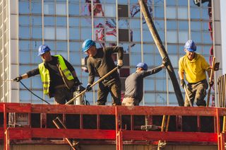 Four construction workers in helmets and safety gear work on a building site, surrounded by scaffolding, with a glass building in the background.
