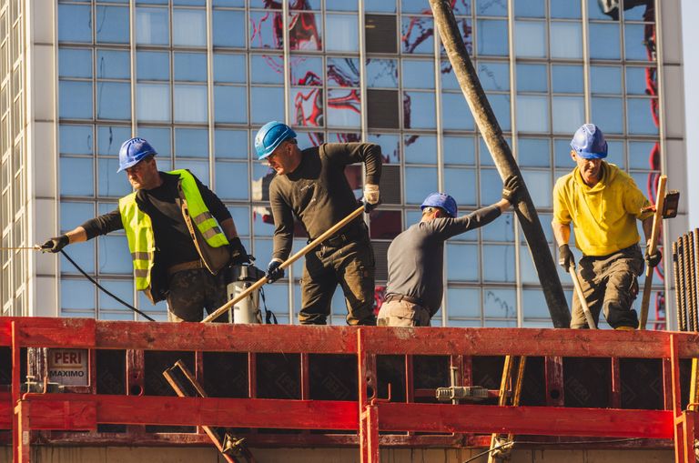Four construction workers in helmets and safety gear work on a building site, surrounded by scaffolding, with a glass building in the background.