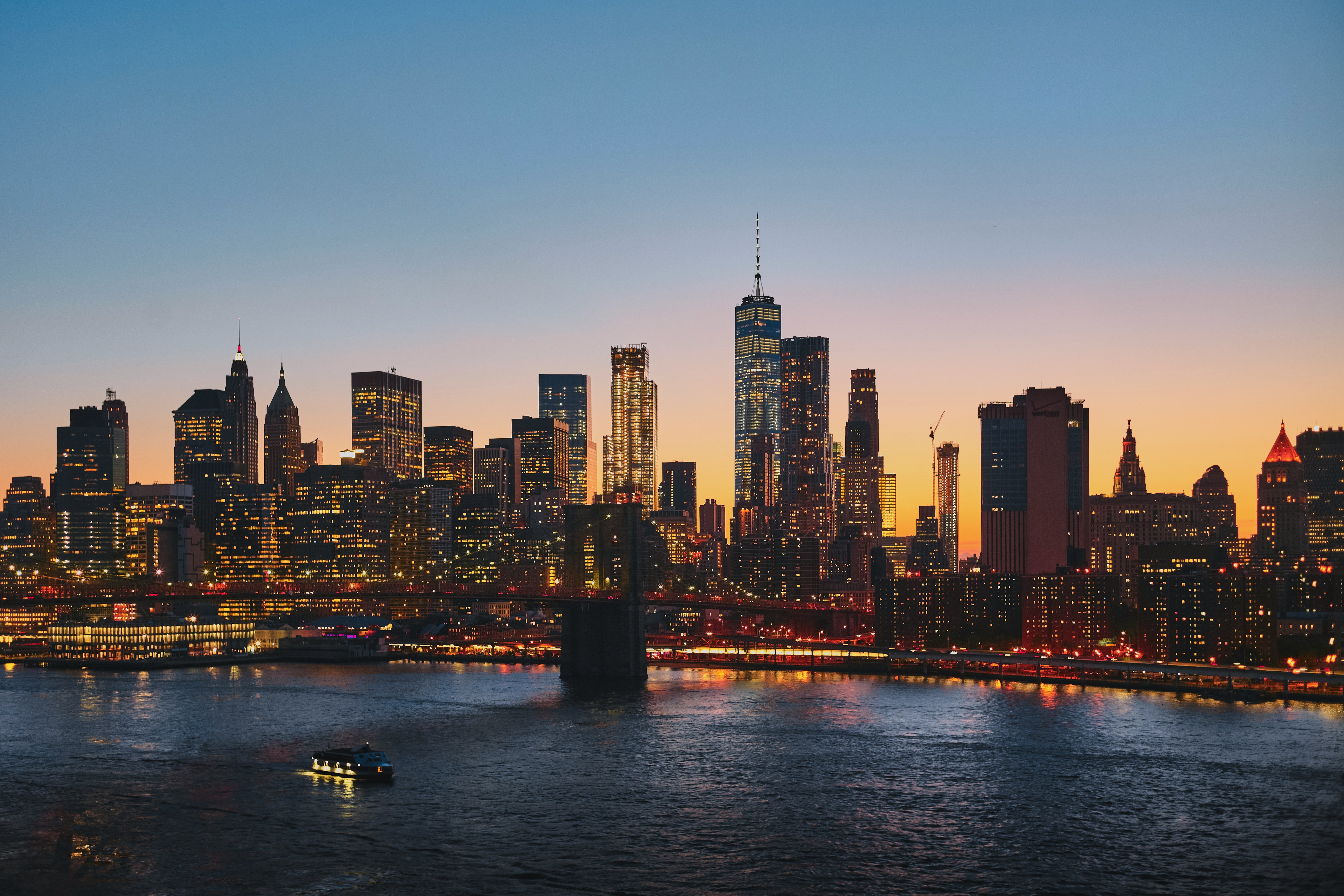 City skyline at sunset with illuminated skyscrapers, a river below reflecting lights, and a small boat in the foreground.