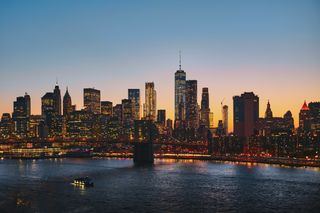 City skyline at sunset with illuminated skyscrapers, a river below reflecting lights, and a small boat in the foreground.