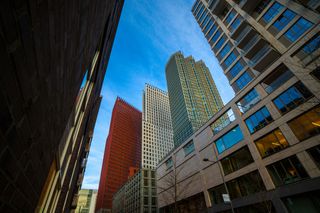 Tall modern skyscrapers against a blue sky, viewed from a low angle between two buildings, creating a dynamic urban perspective.