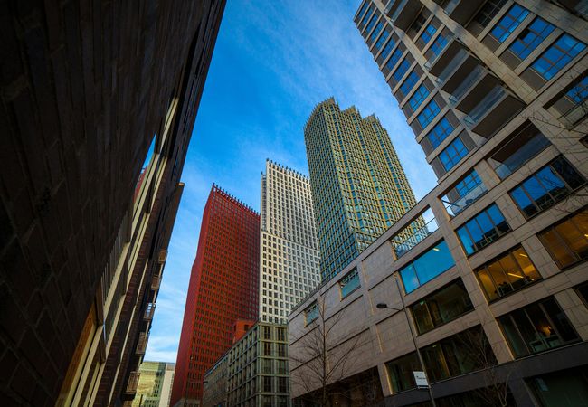 Tall modern skyscrapers against a blue sky, viewed from a low angle between two buildings, creating a dynamic urban perspective.