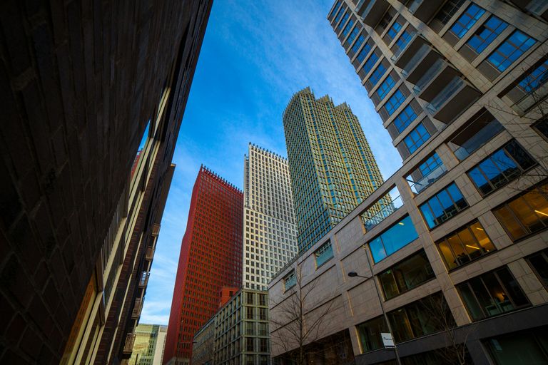 Tall modern skyscrapers against a blue sky, viewed from a low angle between two buildings, creating a dynamic urban perspective.