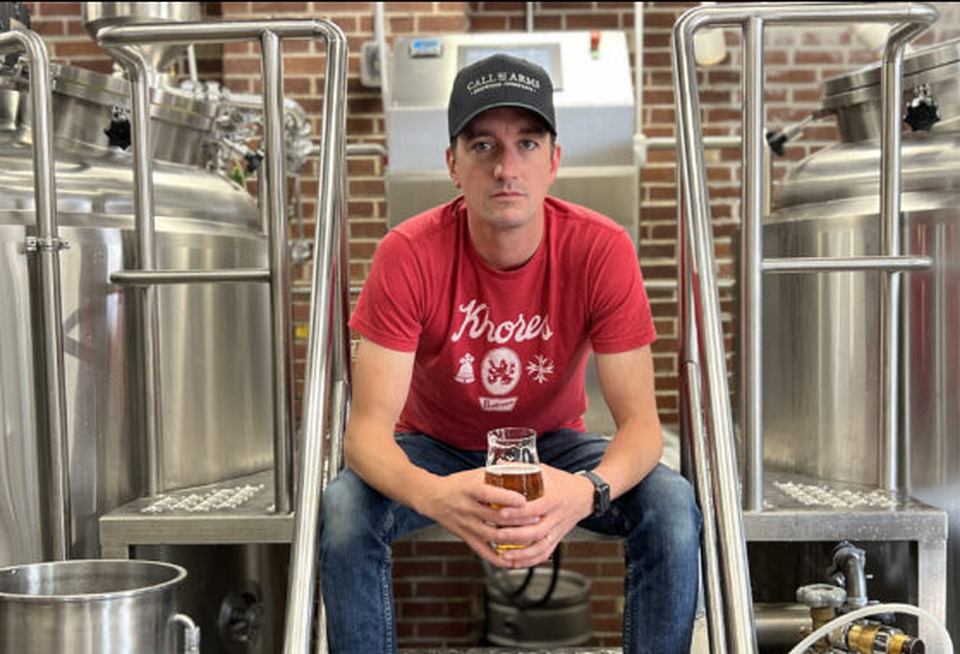 A professional portrait photo of Chris Bell, the owner of Call To Arms Brewing Co., wearing a red shirt and black hat in his brewery.