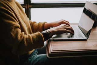 Person in a brown sweater typing on a laptop at a wooden desk.