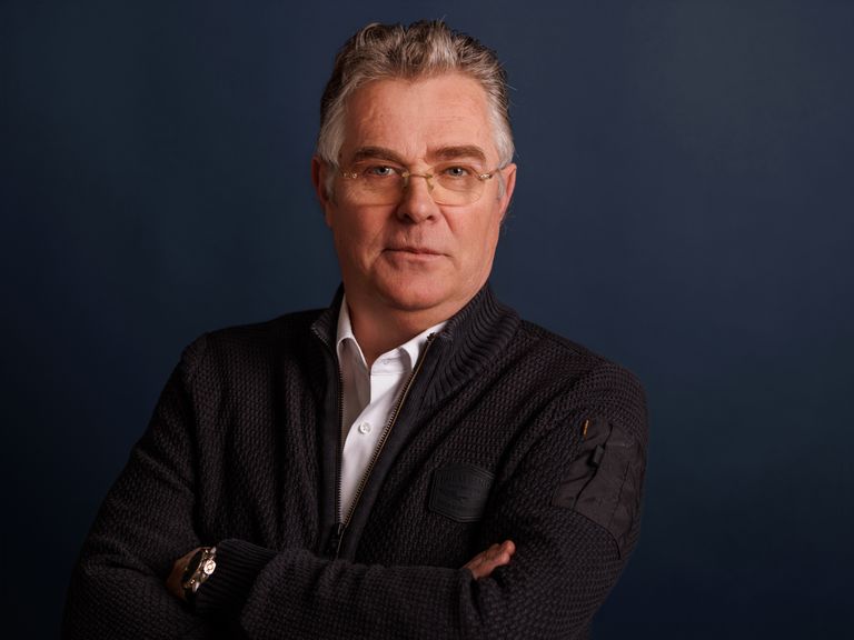 Older man with glasses, wearing a dark sweater and white shirt, stands with arms crossed against a dark blue background.