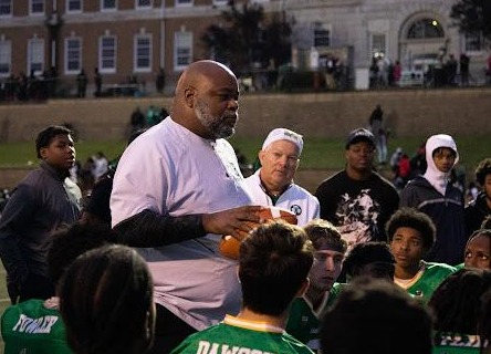 A coach talks to a group of young football players wearing green jerseys on a field, with a large building visible in the background.