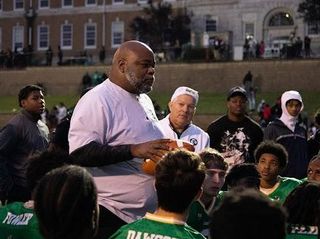 A coach talks to a group of young football players wearing green jerseys on a field, with a large building visible in the background.