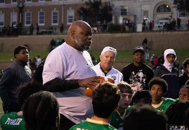A coach talks to a group of young football players wearing green jerseys on a field, with a large building visible in the background.
