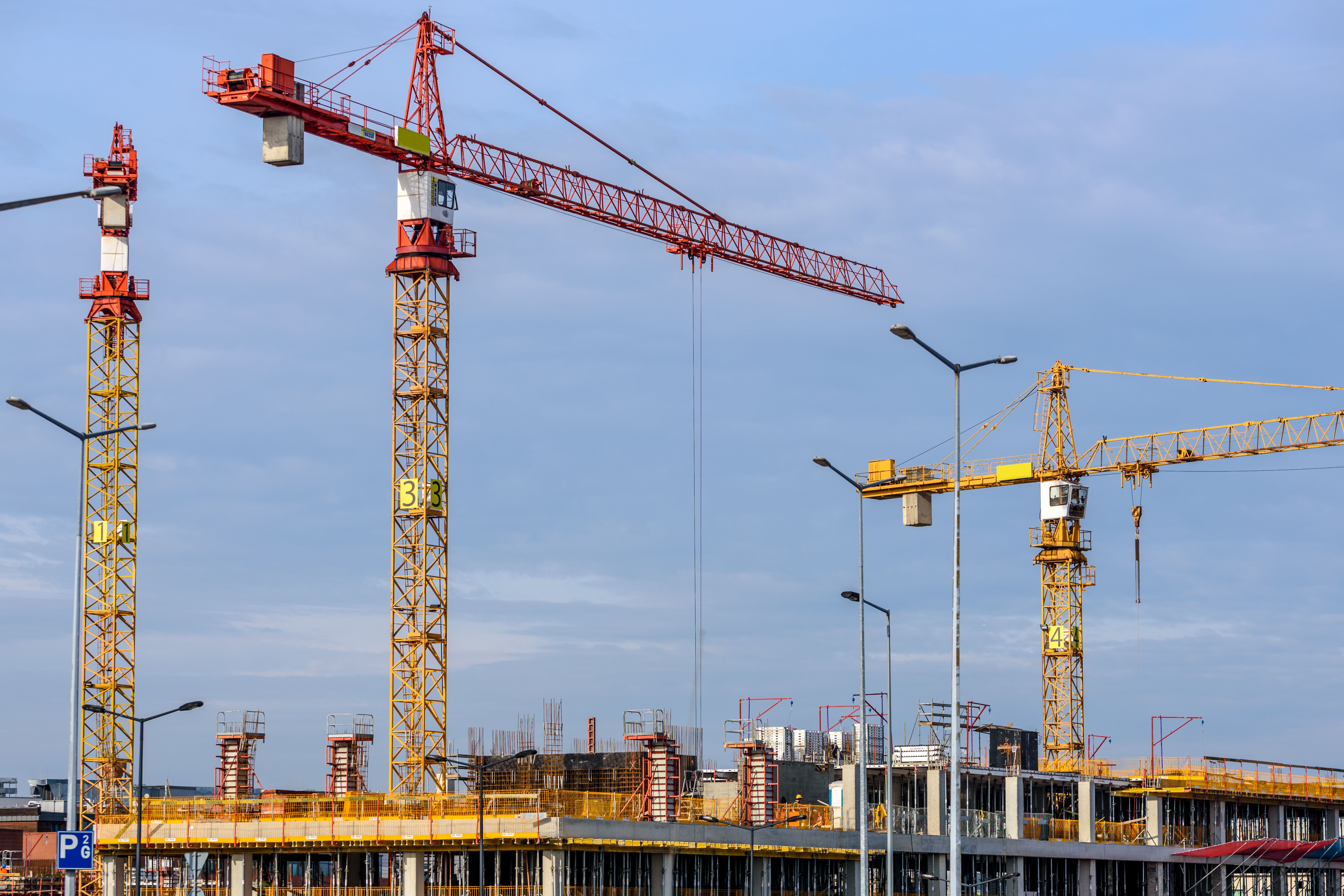 Construction site with three large cranes towering over a partially built structure, under a clear blue sky.