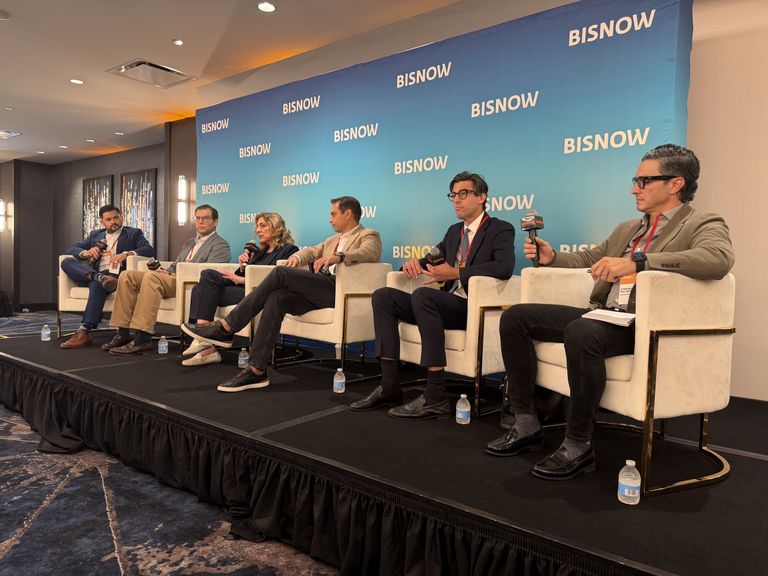 A panel of six people sitting on stage at a conference, each holding microphones, with a "BISNOW" backdrop and bottled water at their feet.