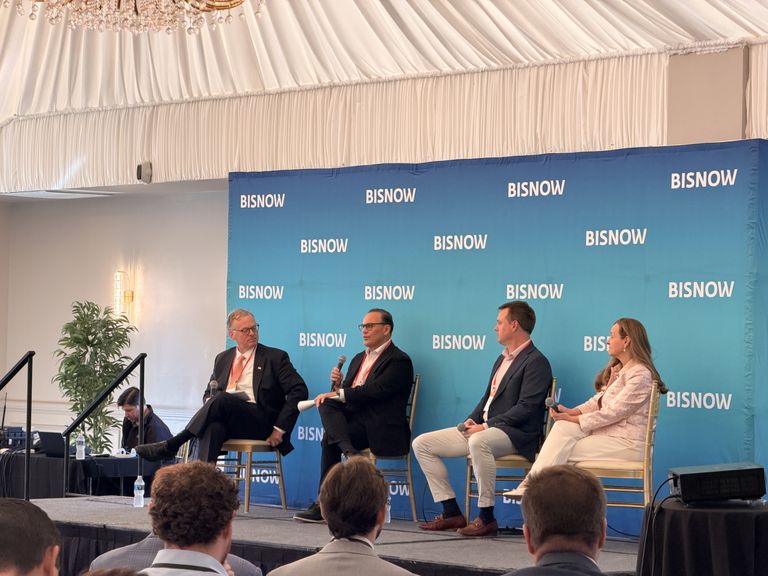 Four people seated on a panel discussion stage with a blue "BISNOW" backdrop, under a draped ceiling and chandelier.