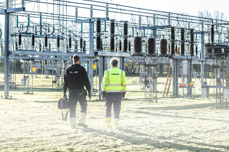 Two workers wearing safety gear walk through an outdoor electrical substation, surrounded by high-voltage equipment under a clear sky.