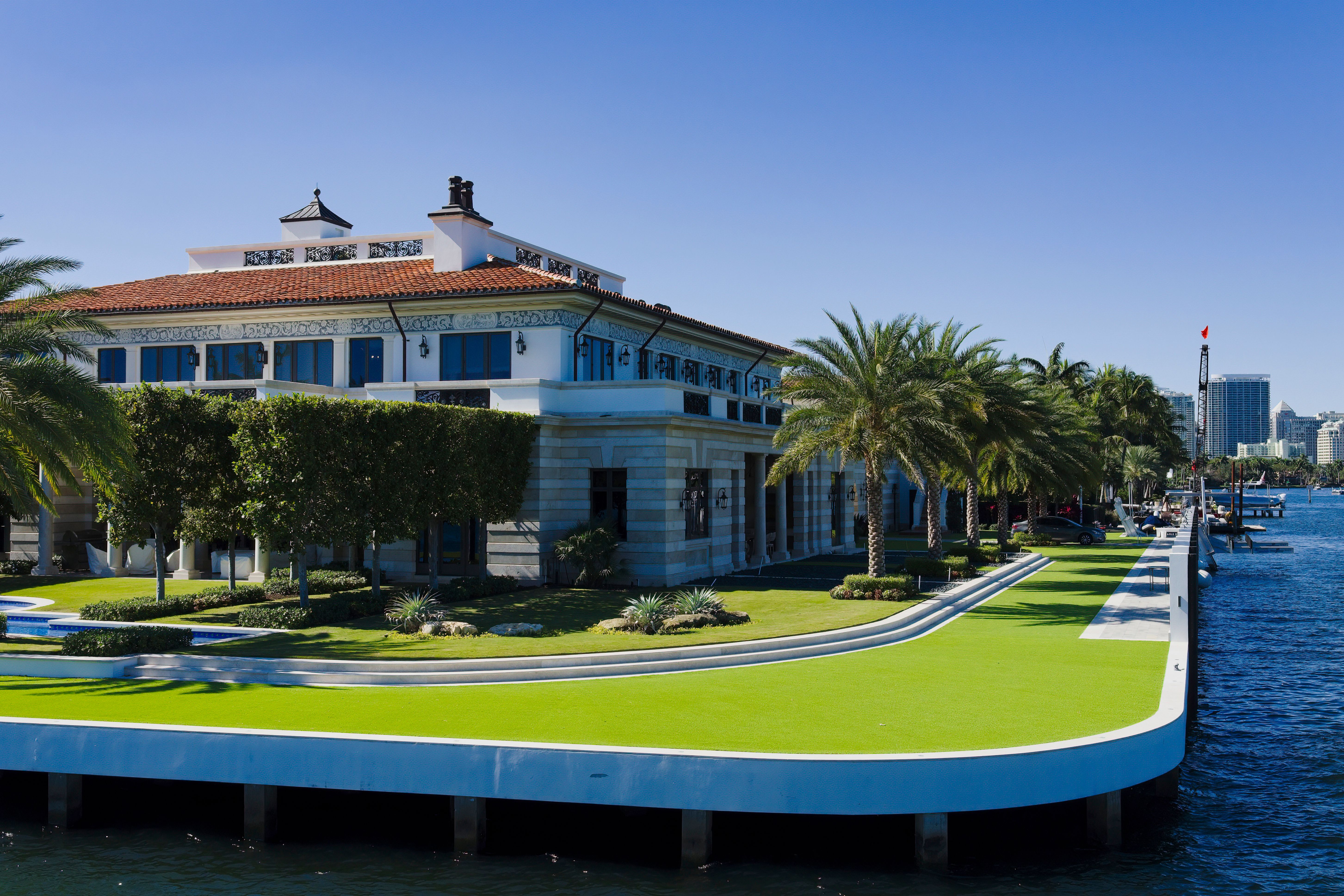 Waterfront mansion with a red-tiled roof, surrounded by palm trees and a manicured lawn, overlooking a calm body of water under a clear blue sky.