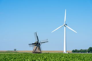 A classic windmill and a modern wind turbine stand in a lush green field under a clear blue sky.