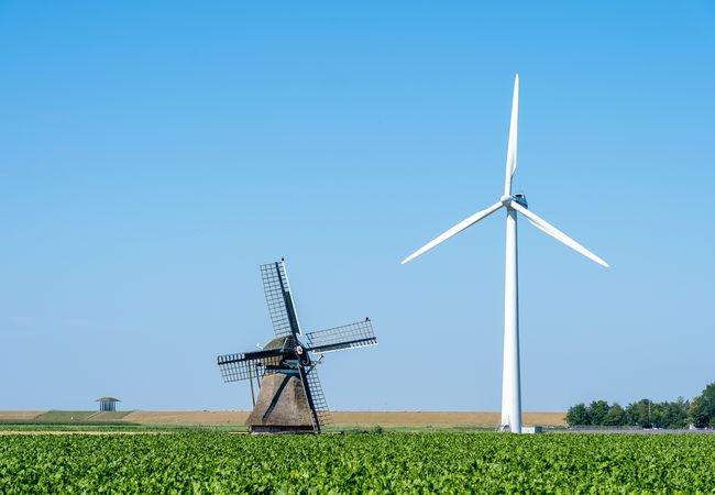 A classic windmill and a modern wind turbine stand in a lush green field under a clear blue sky.
