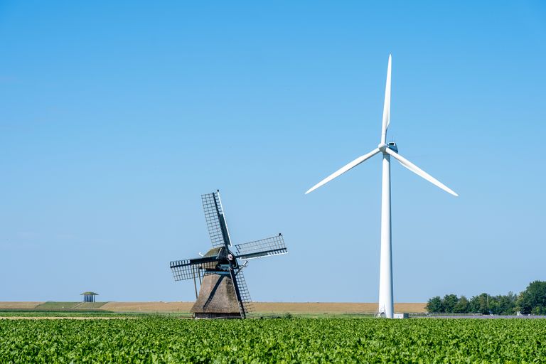 A classic windmill and a modern wind turbine stand in a lush green field under a clear blue sky.